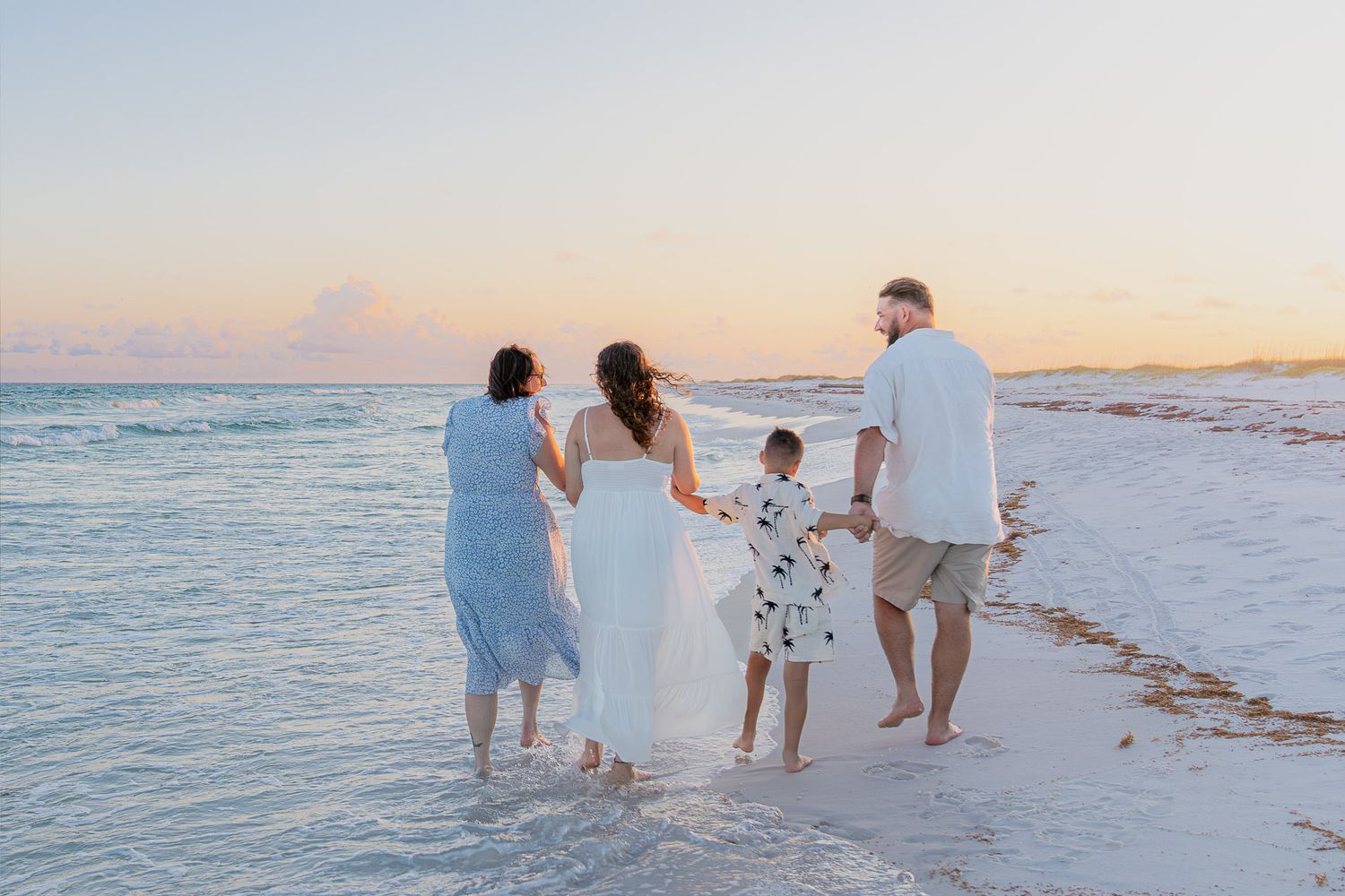 Family of four walking hand-in-hand along a sandy beach at sunset, waves gently lapping at their feet.
