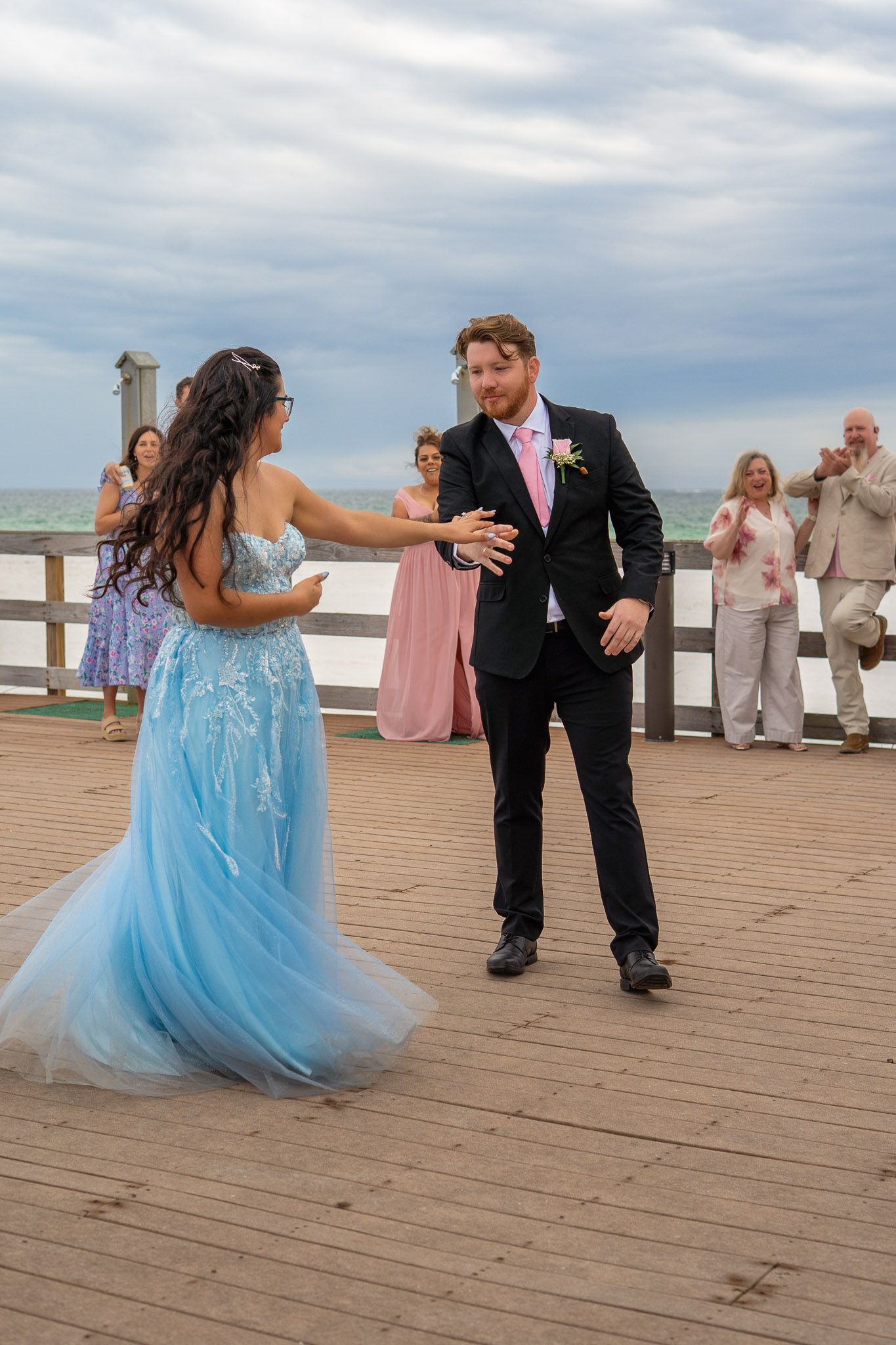 Couple dancing outdoors on wooden deck, woman in blue tulle gown and man in black suit with pink tie.