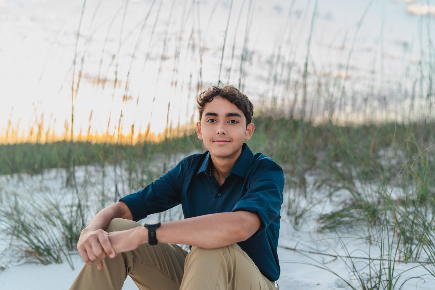 Young man in navy shirt sits on white sand dunes with sea oats at sunset beach.