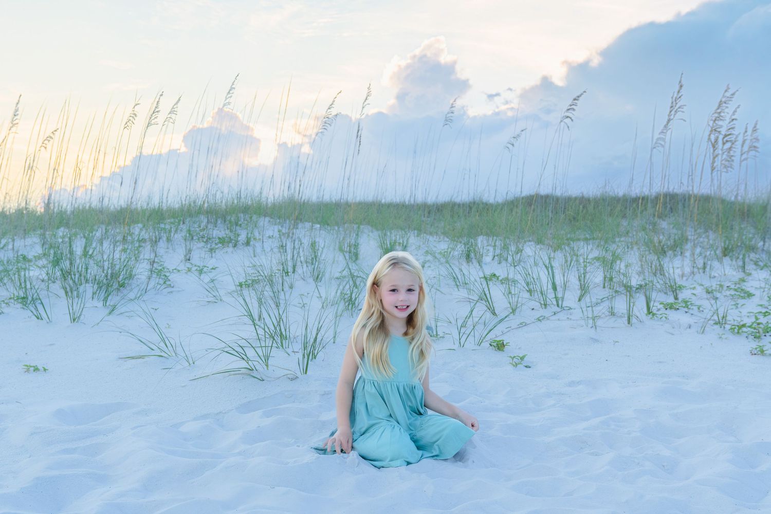 Young blonde girl in teal dress sitting on white sand dunes with sea oats at sunset.