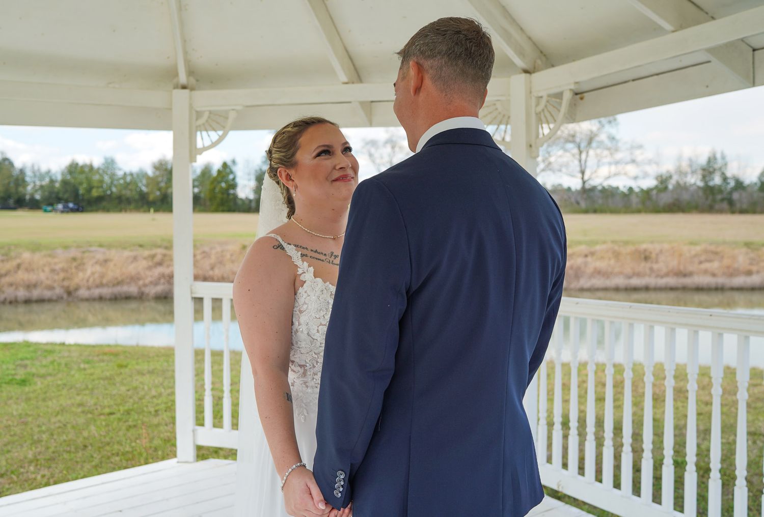Bride in lace wedding gown and groom in navy suit share first look moment in white gazebo.