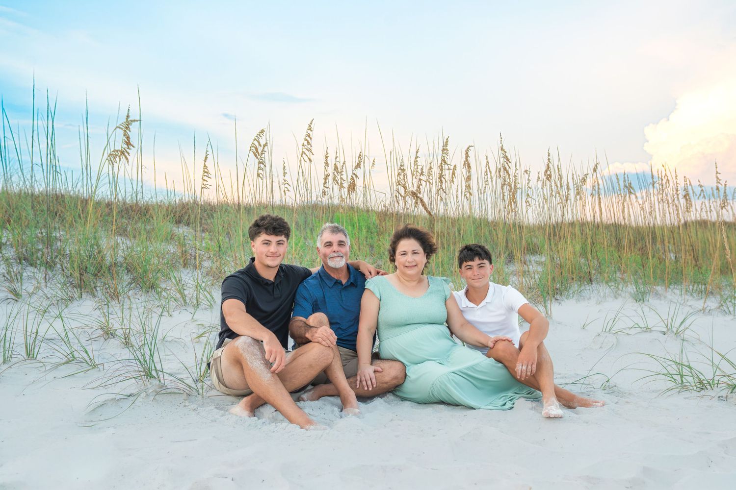 Family of four posing together on white sand beach dunes at sunset with sea oats in background.