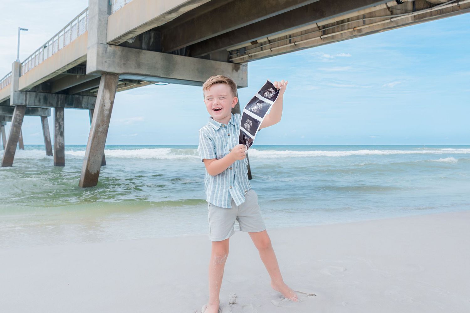 A young boy stands on a beach holding ultrasound photos, smiling joyfully near a pier with ocean waves behind him.