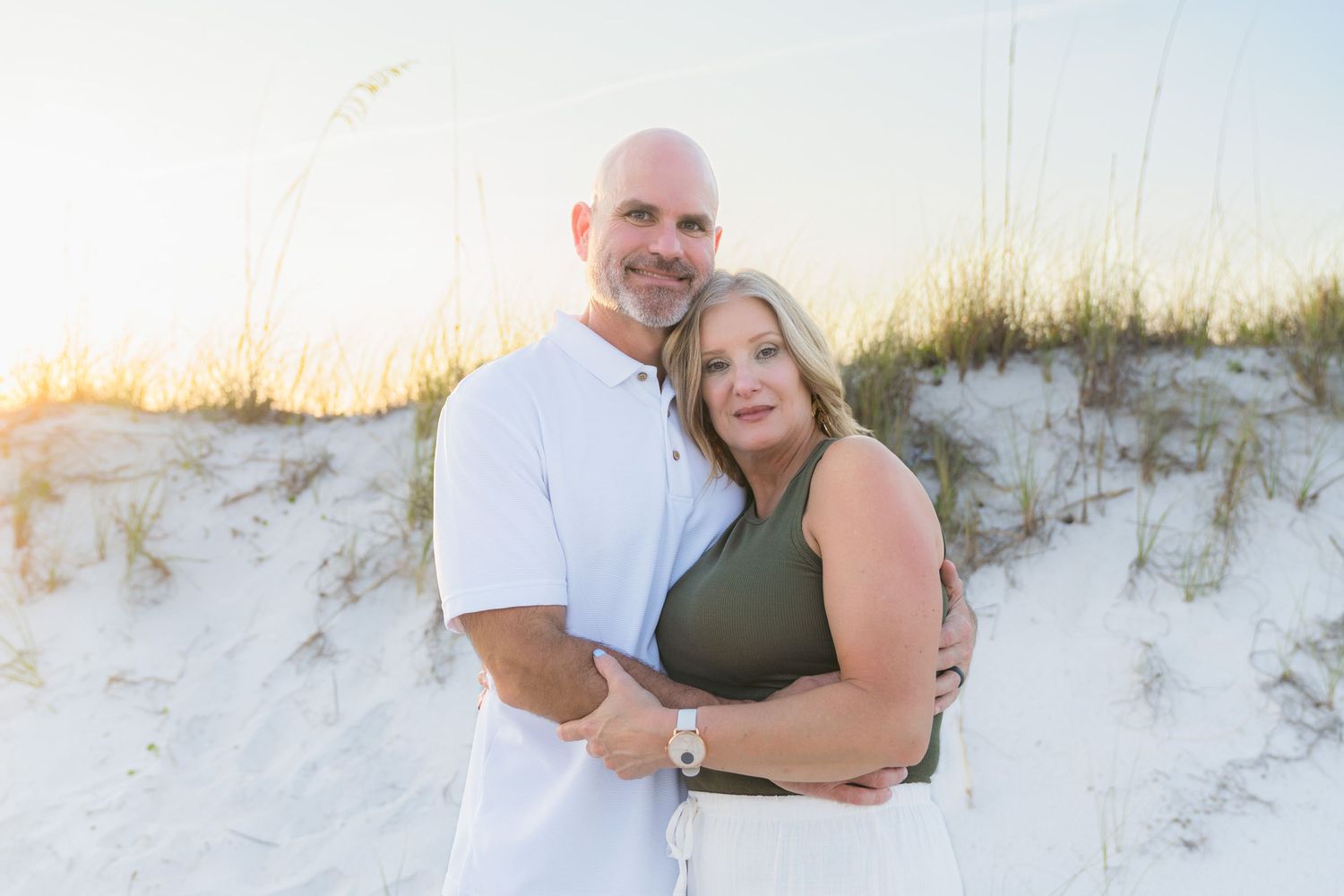 Couple embracing on white sand beach dunes at sunset, man in white shirt and woman in olive top.