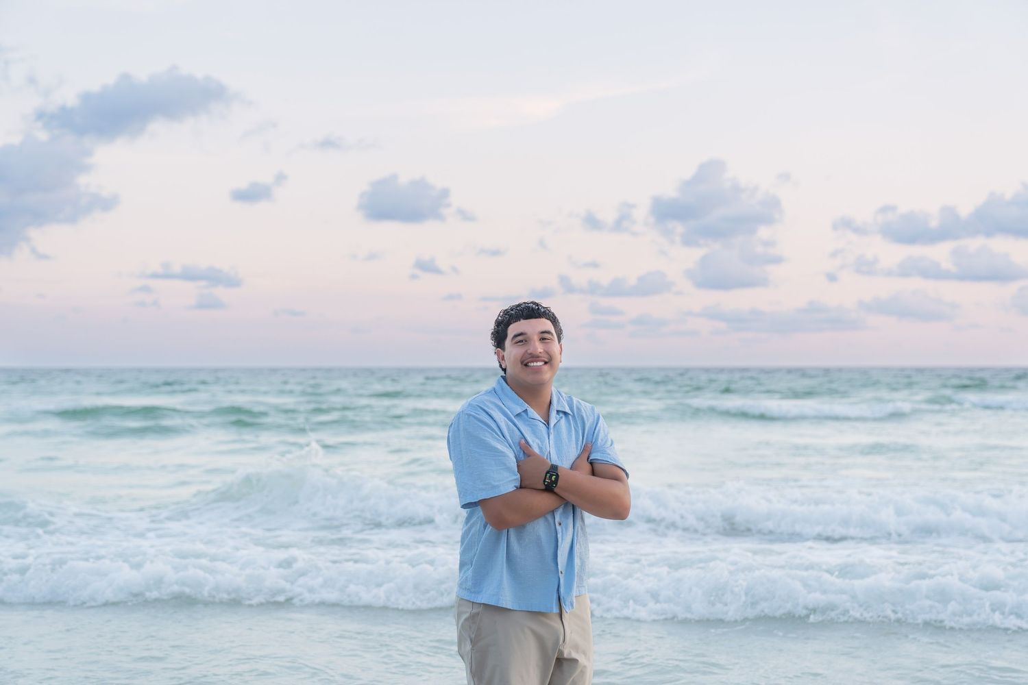 Man smiling with arms crossed on a beach at sunset, wearing a light blue shirt with ocean waves behind him.