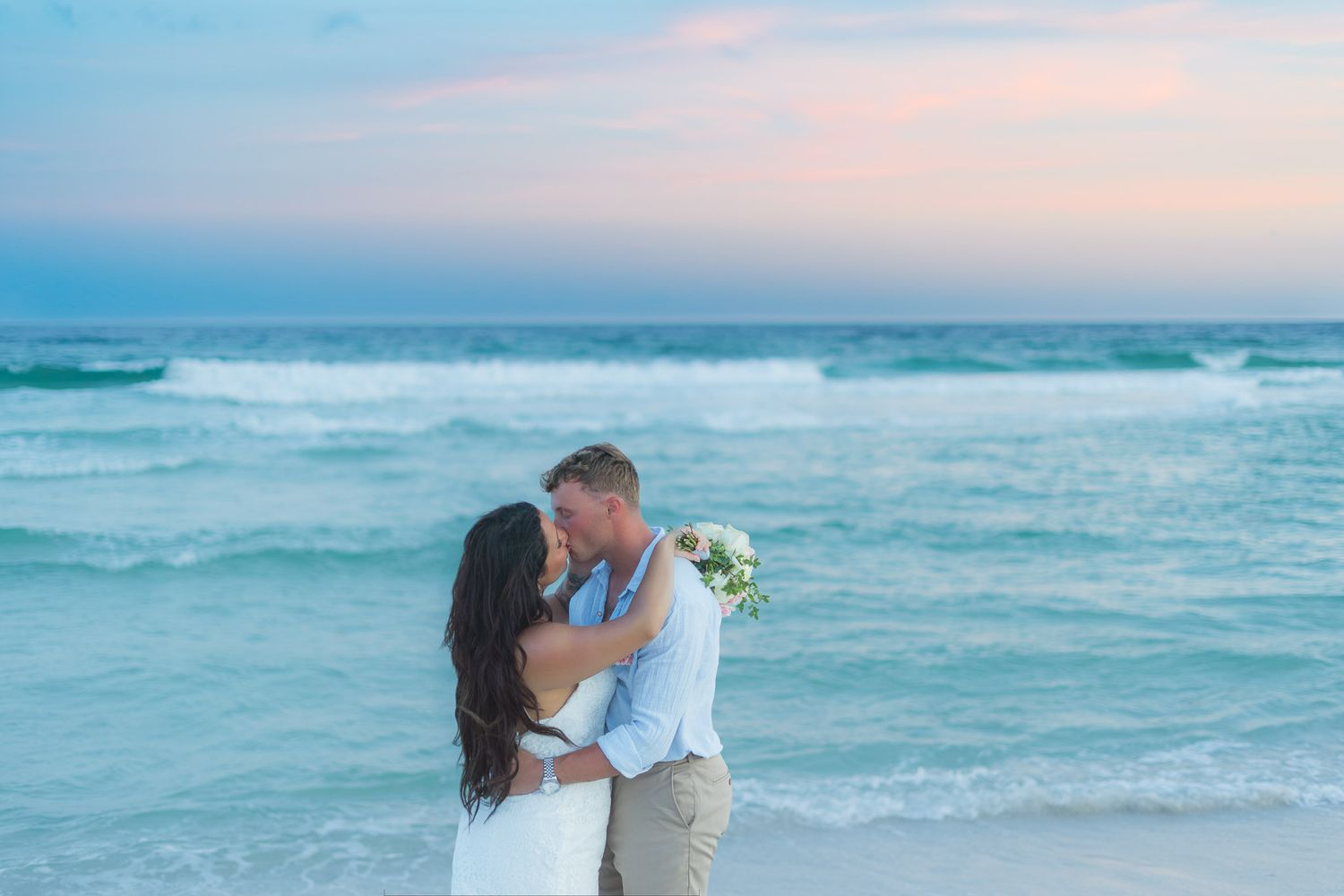 Newlyweds share first kiss on beach at sunset with turquoise ocean waves and pastel sky in the background.