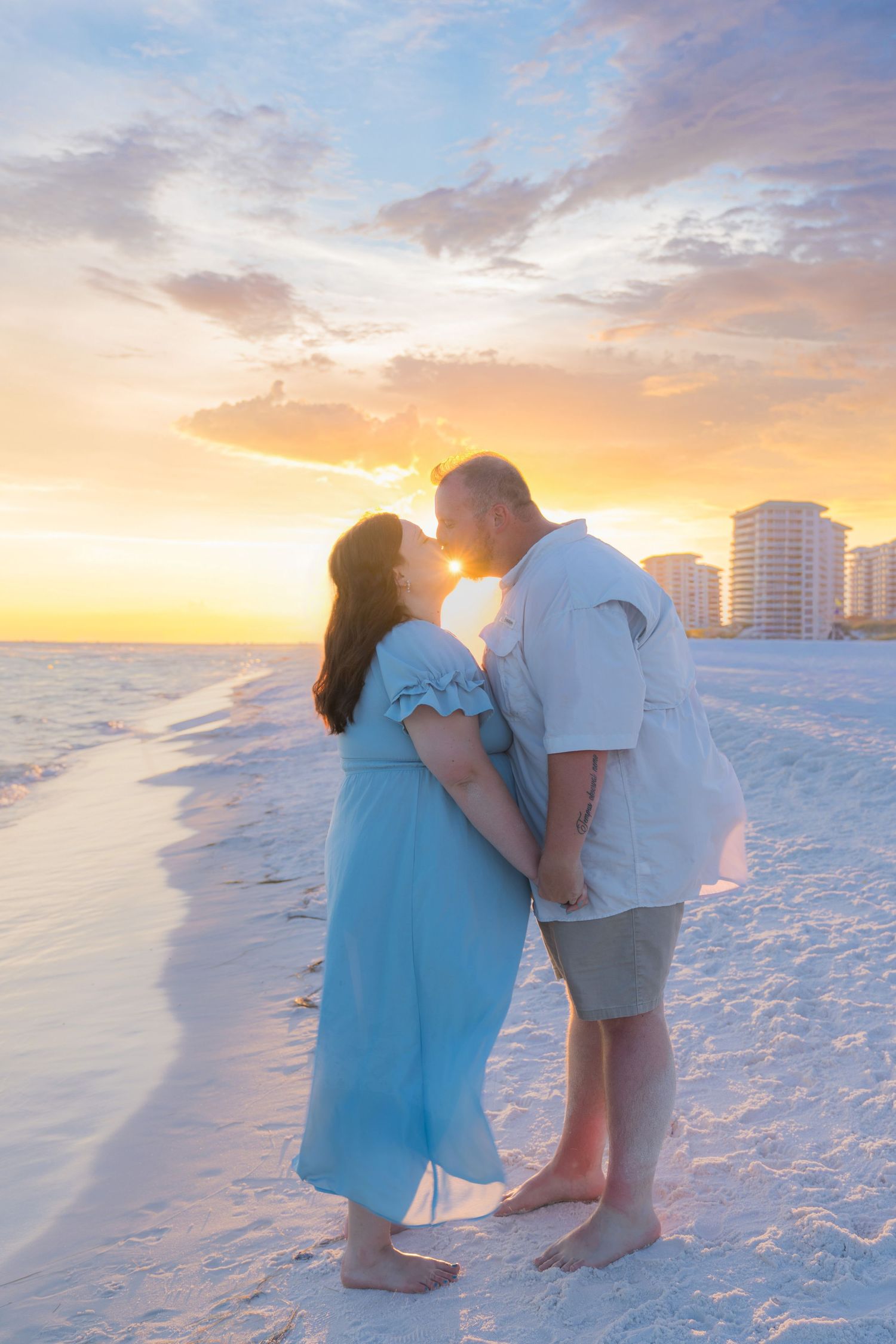 Couple kissing on white sand beach at sunset, woman in blue dress, man in white shirt, colorful sky backdrop.