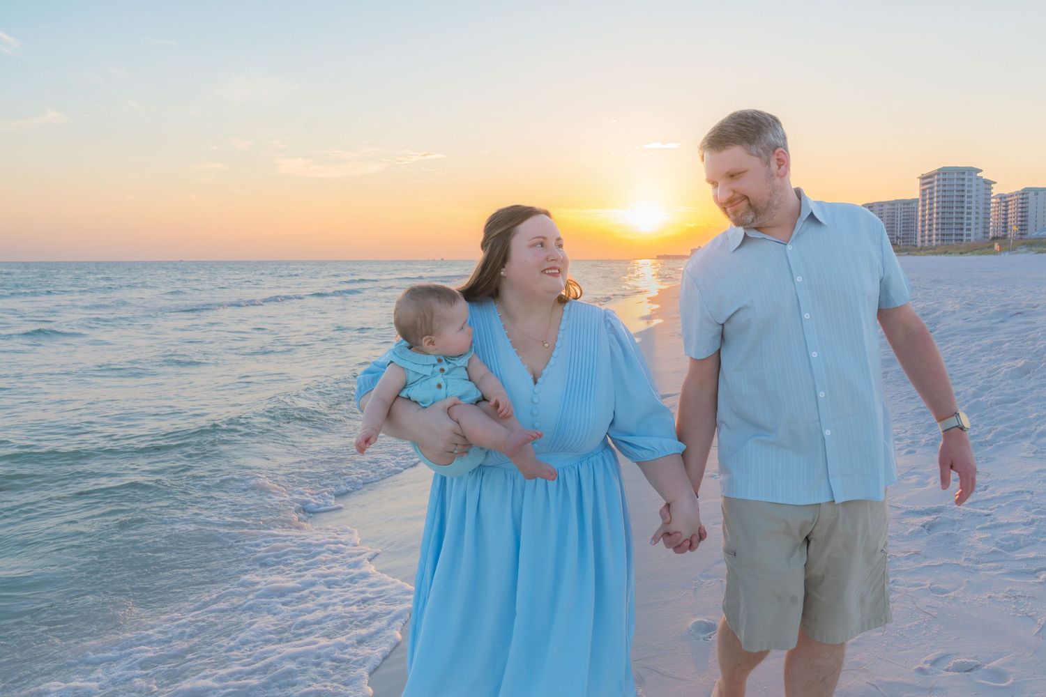 Family of three walking on beach at sunset, parents holding hands with mother carrying baby in blue dress.