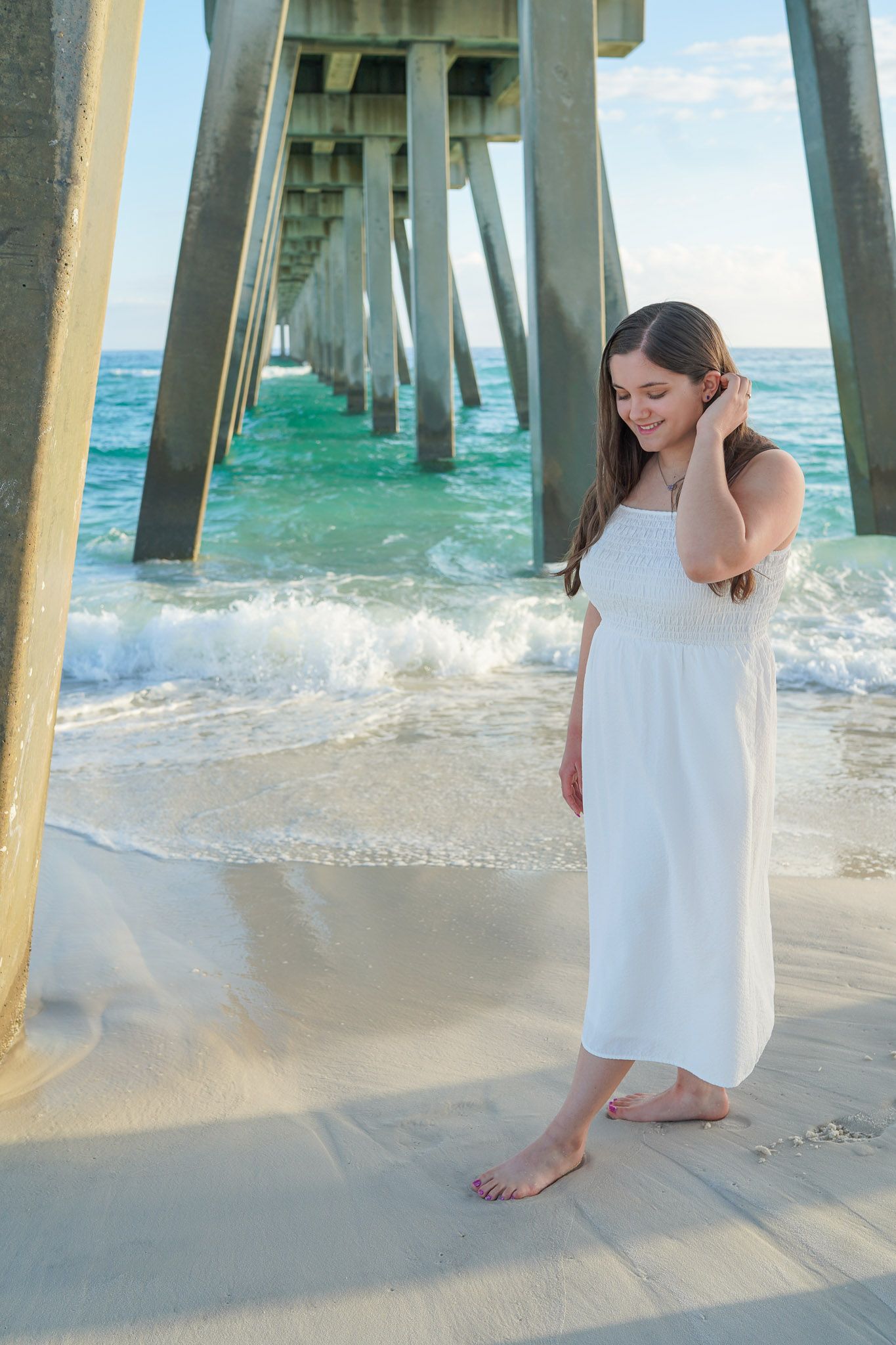 Young woman in white dress standing barefoot on sandy beach under a wooden pier with turquoise ocean waves.