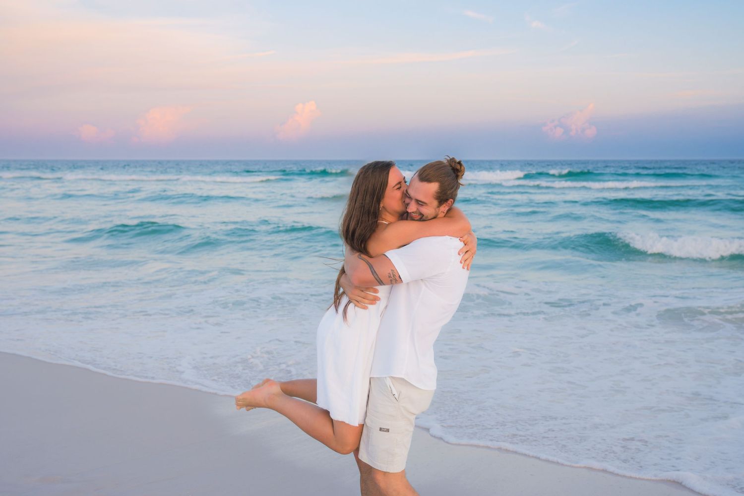 A couple embraces romantically on a tropical beach at sunset, both wearing white outfits with turquoise waves behind them.