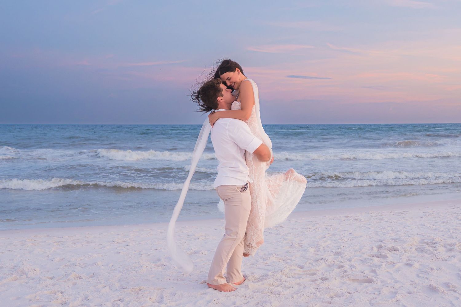 Groom lifts bride in white wedding dress on sandy beach at pastel sunset by ocean waves.
