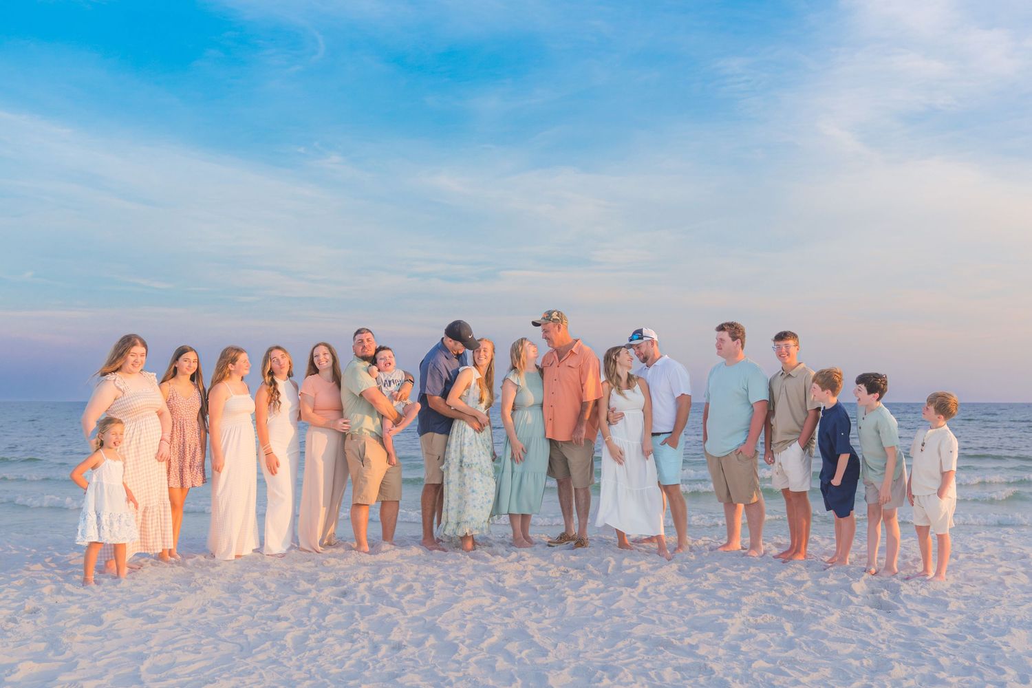 Large family group posing together on a sandy beach at sunset in coordinated pastel outfits.