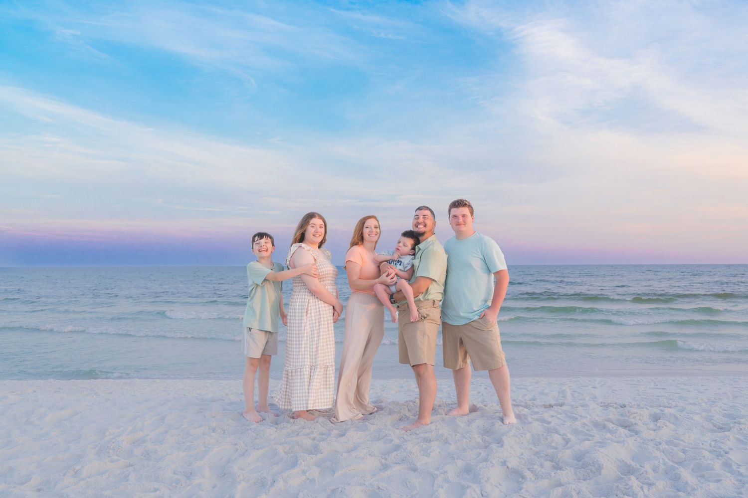 Family of five poses on a sandy beach at sunset with pastel skies and calm ocean waters in the background.