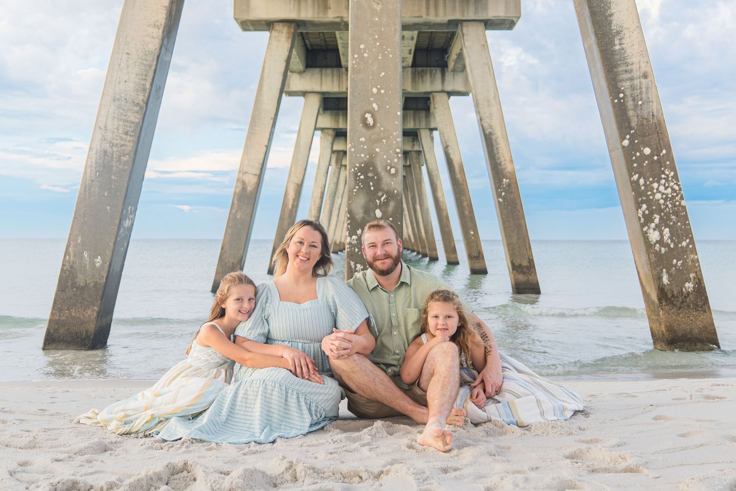 Family of four smiling together on sandy beach under a wooden pier, dressed in soft blue and green tones.