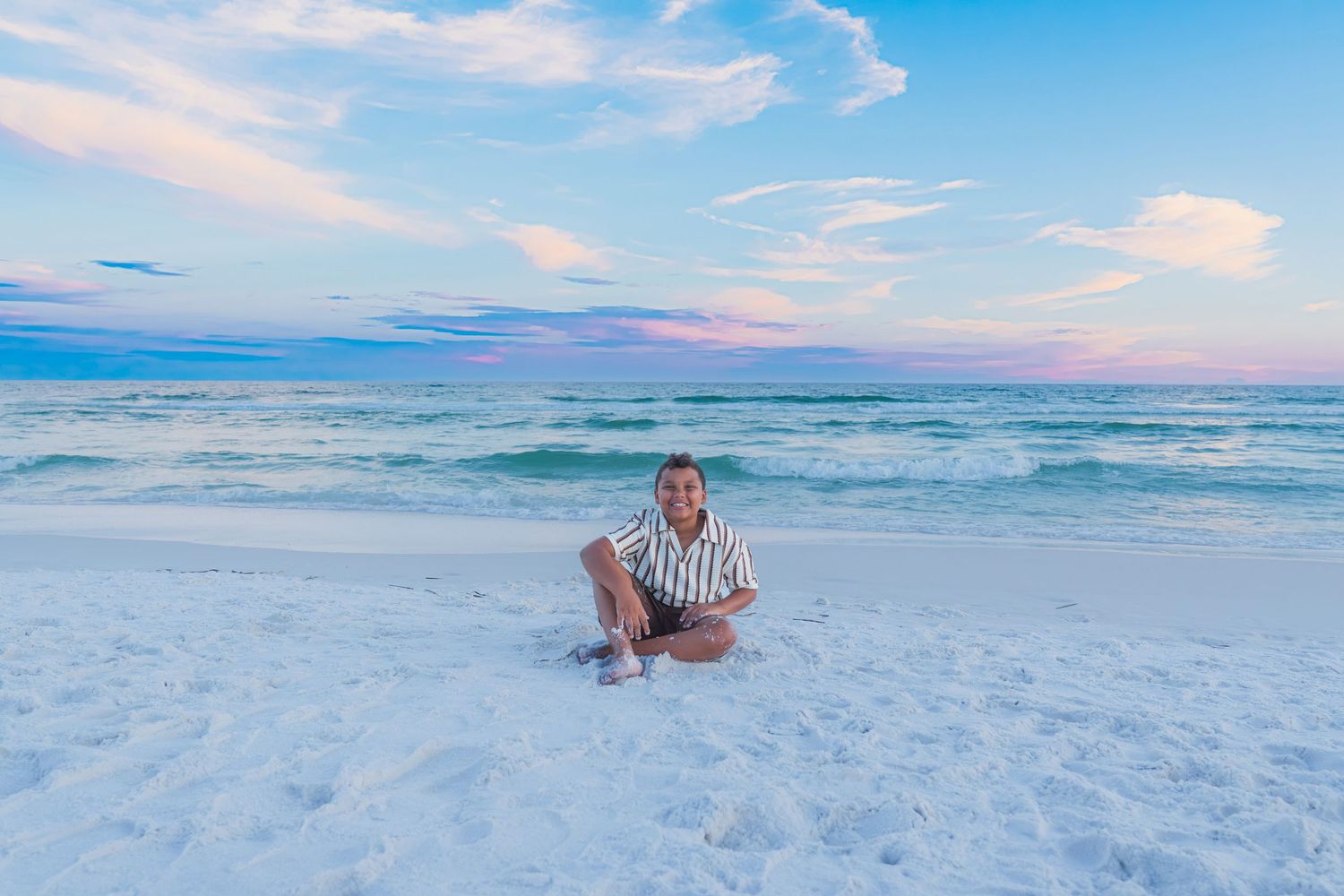 Person sitting cross-legged on white sand beach at sunset with calm turquoise ocean waves and pastel sky.