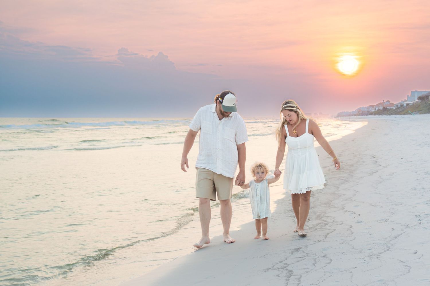 Family of three walking hand-in-hand on a sandy beach at sunset, dressed in white and khaki.