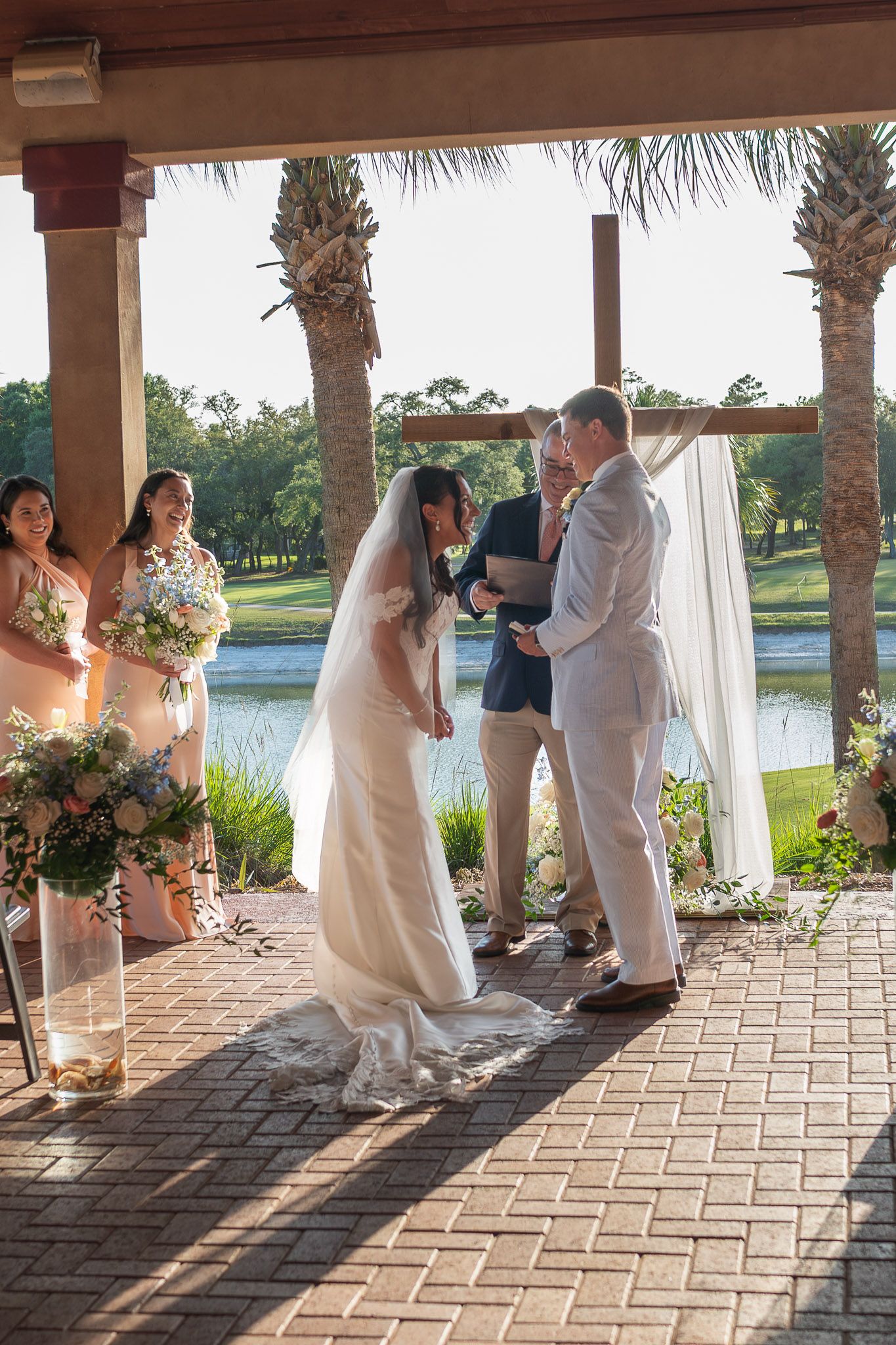 Bride and groom exchanging vows at outdoor wedding ceremony under pergola with palm trees and pond backdrop.