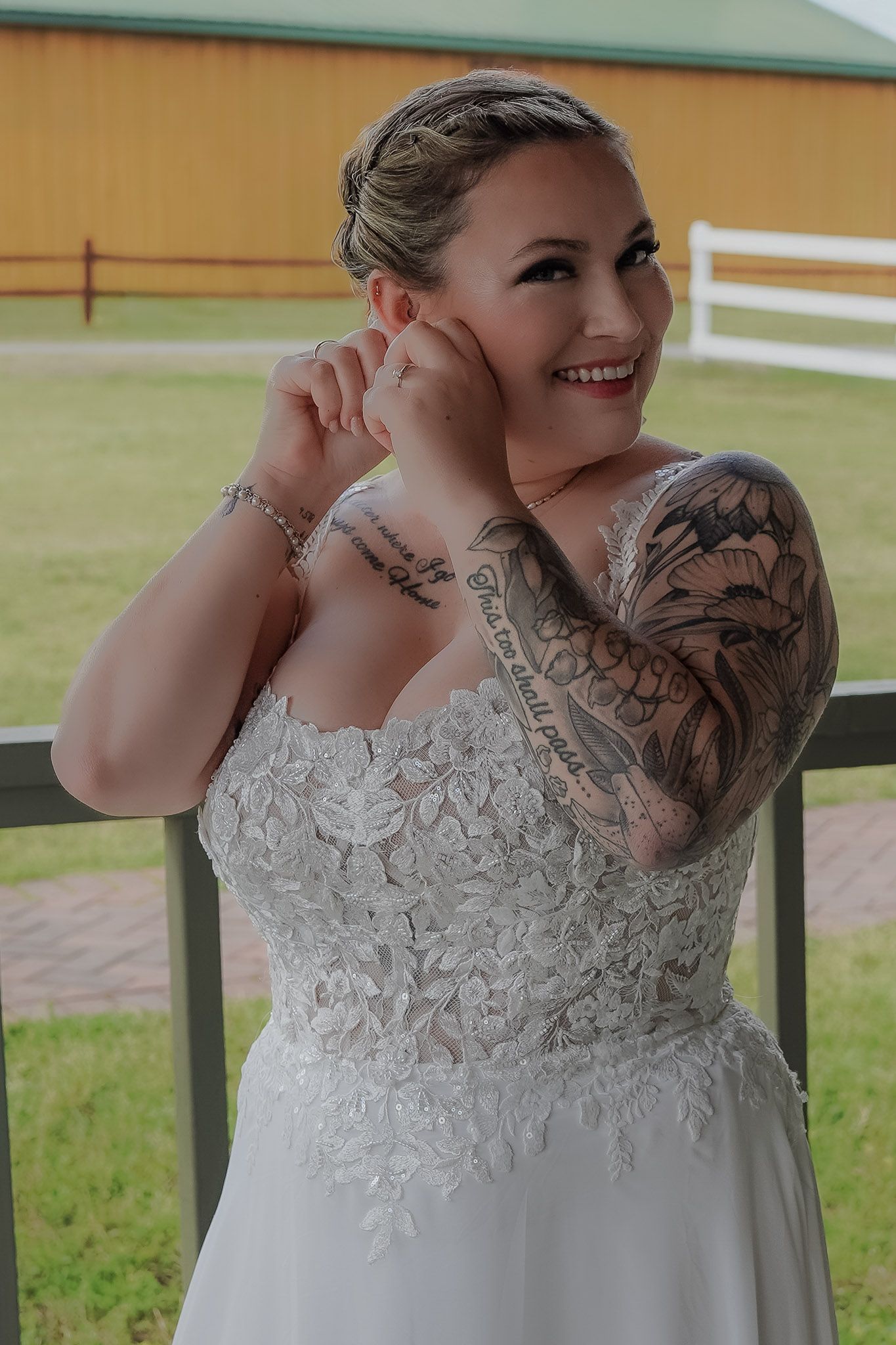 Smiling bride in lace wedding gown with sleeve tattoos poses outdoors near a barn fence.