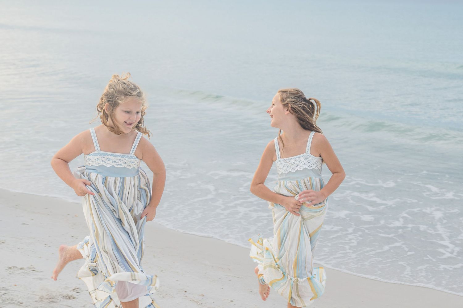 Two girls in matching striped dresses laughing and running along a sunny beach shoreline.
