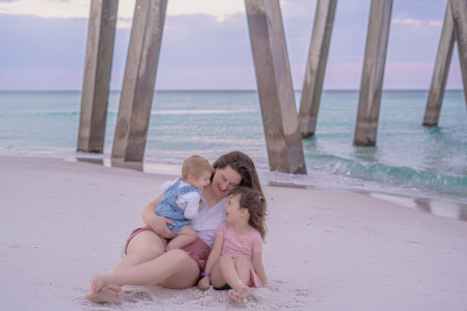 A mother sits on the beach with her two young children near a wooden pier at sunset.