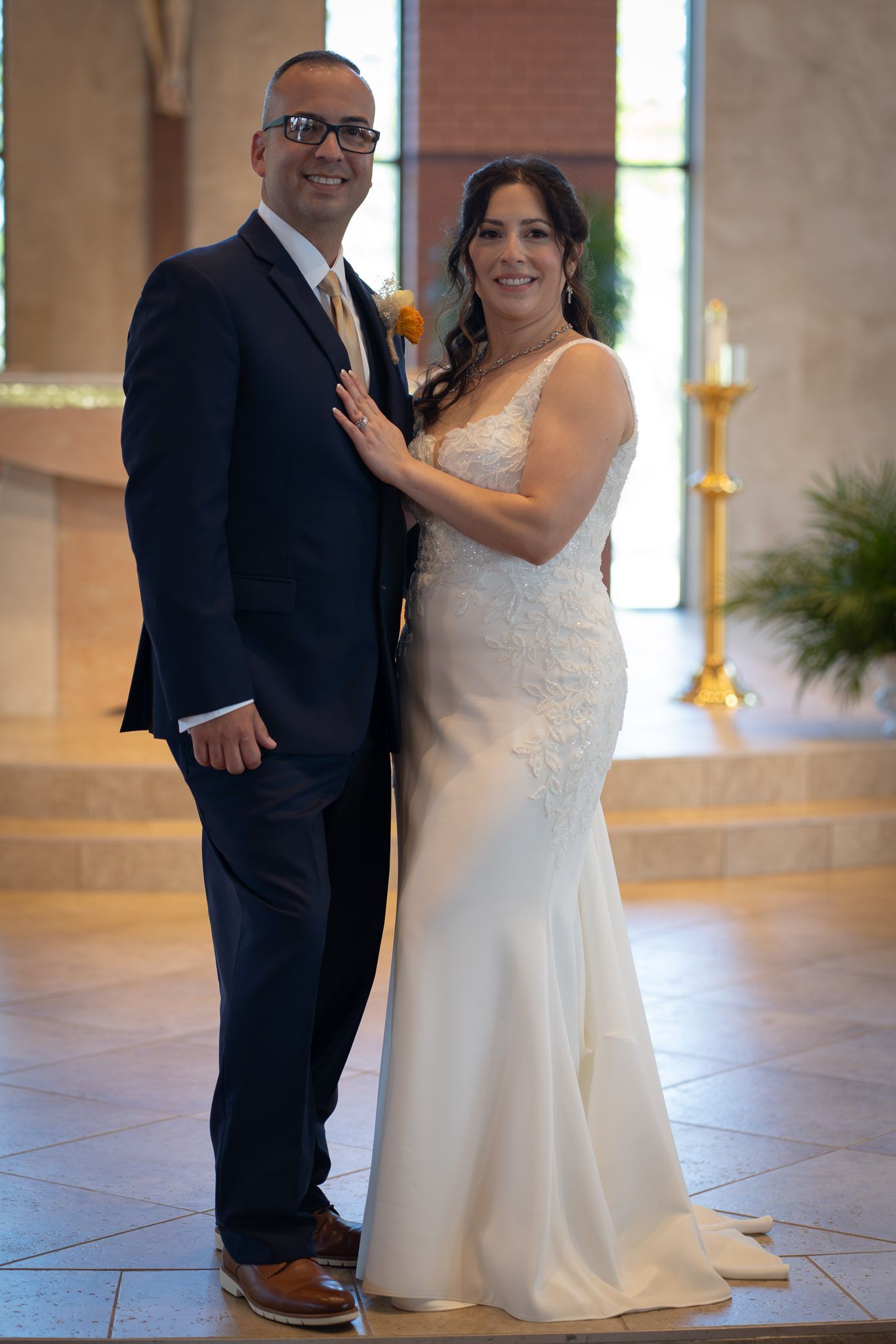 Bride and groom pose together in a church, she in a white lace gown and he in a navy suit with gold boutonniere.