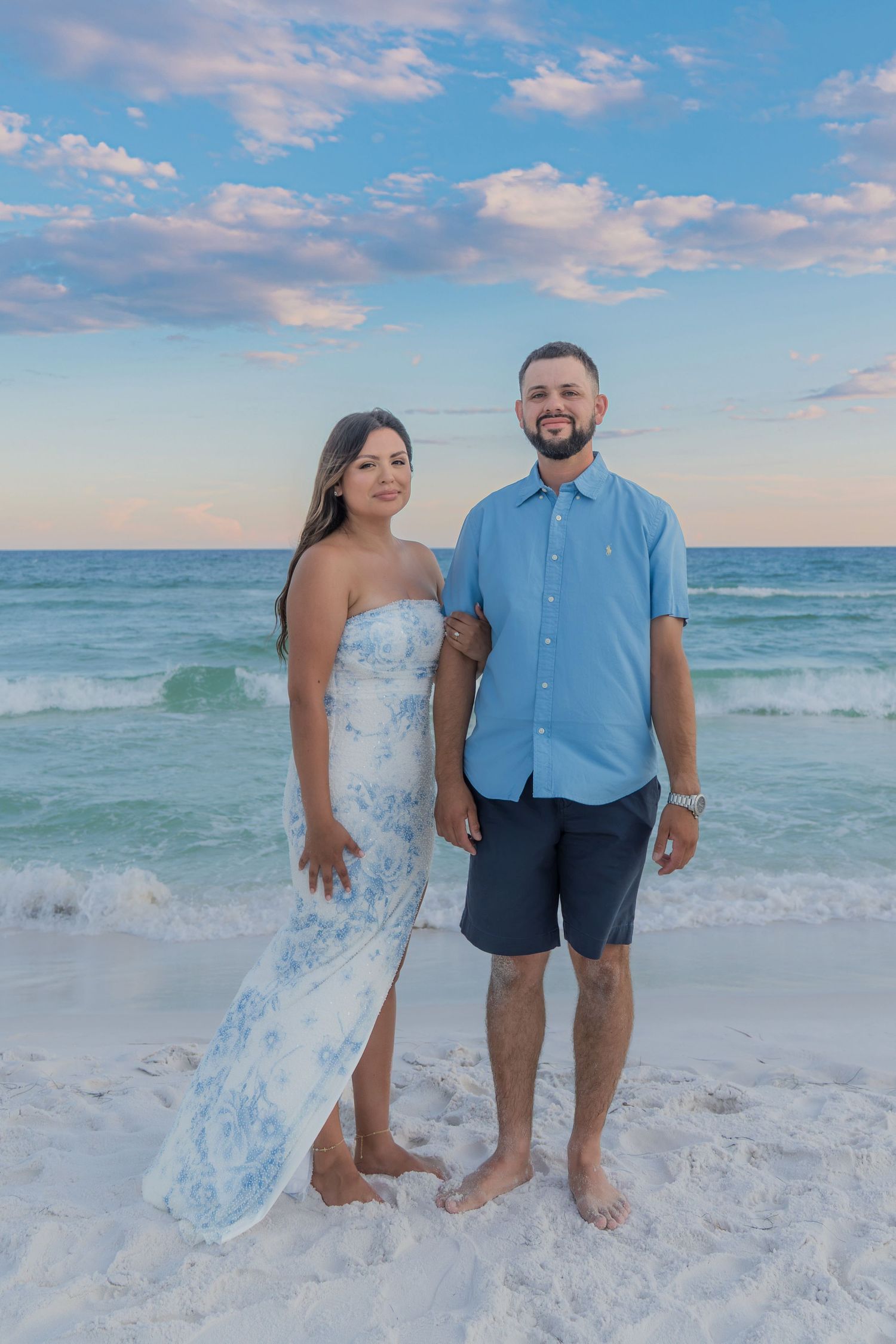 A couple smiling together on a sandy beach with turquoise ocean waves and a blue cloudy sky behind them.
