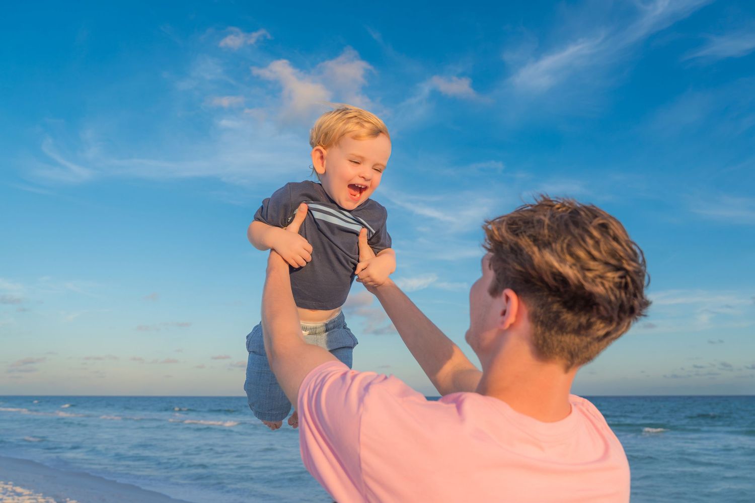 Father joyfully lifting laughing toddler son high in the air on a sunny beach with blue sky and ocean waves.