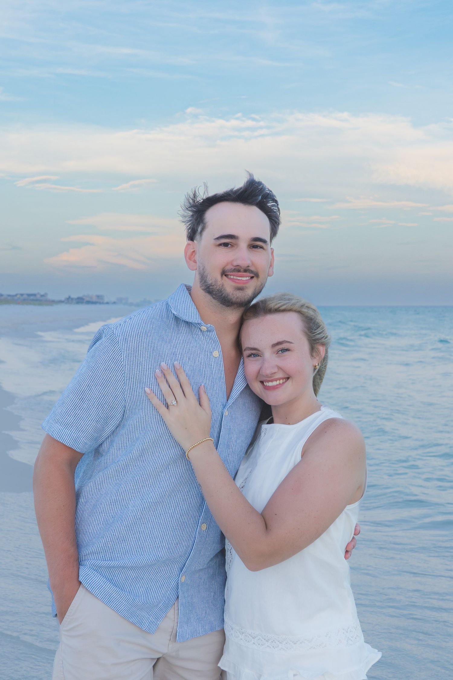 A smiling couple embraces on a sandy beach at sunset, woman wearing white dress showing engagement ring.