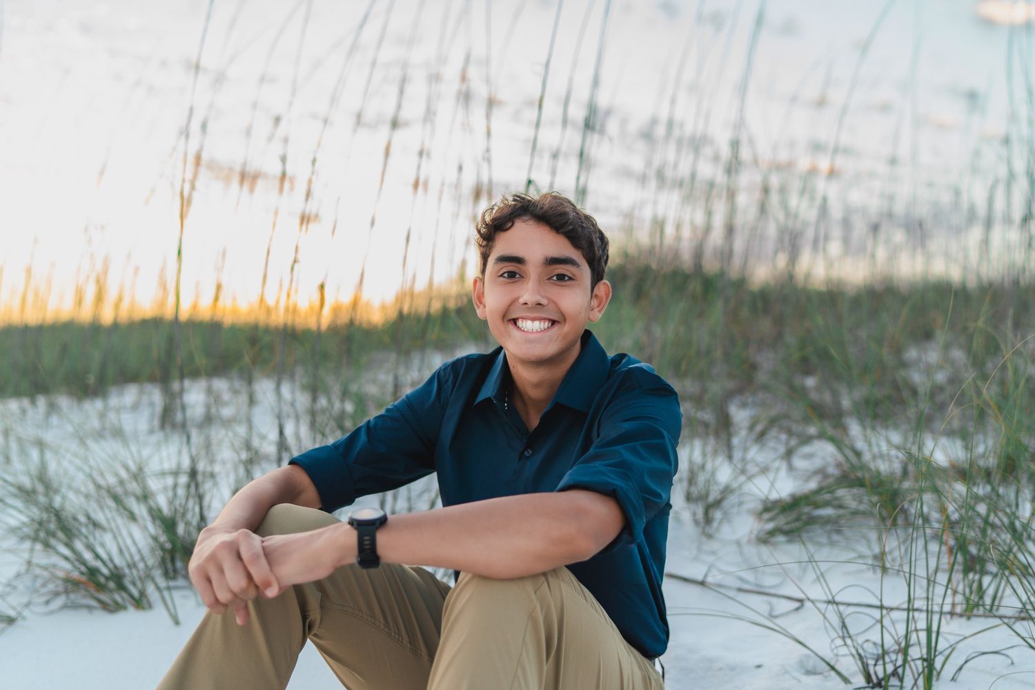 Smiling young man sitting on white sand dunes with sea oats at sunset beach.