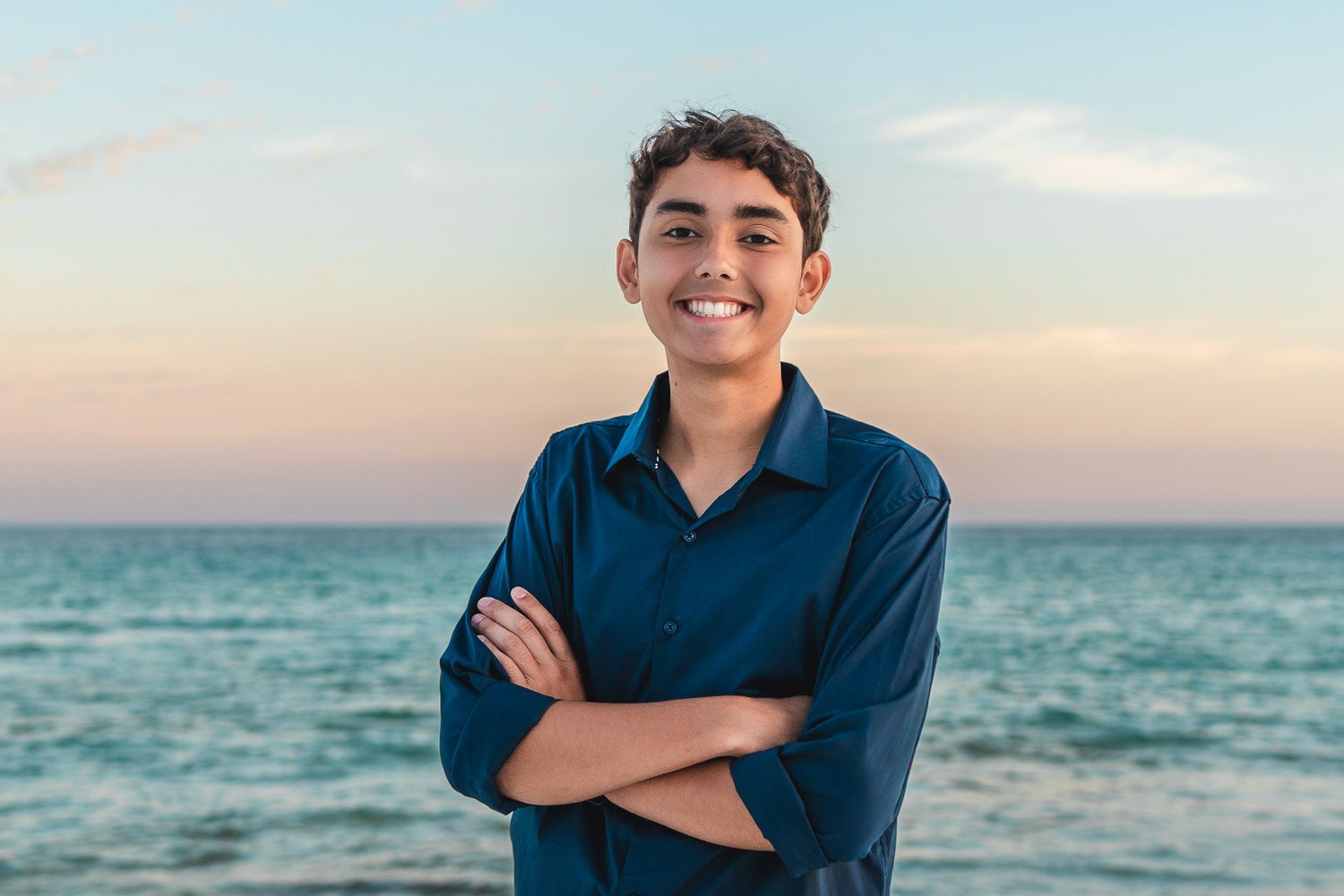 Smiling teenage boy with arms crossed standing on a beach at sunset wearing a dark blue shirt.
