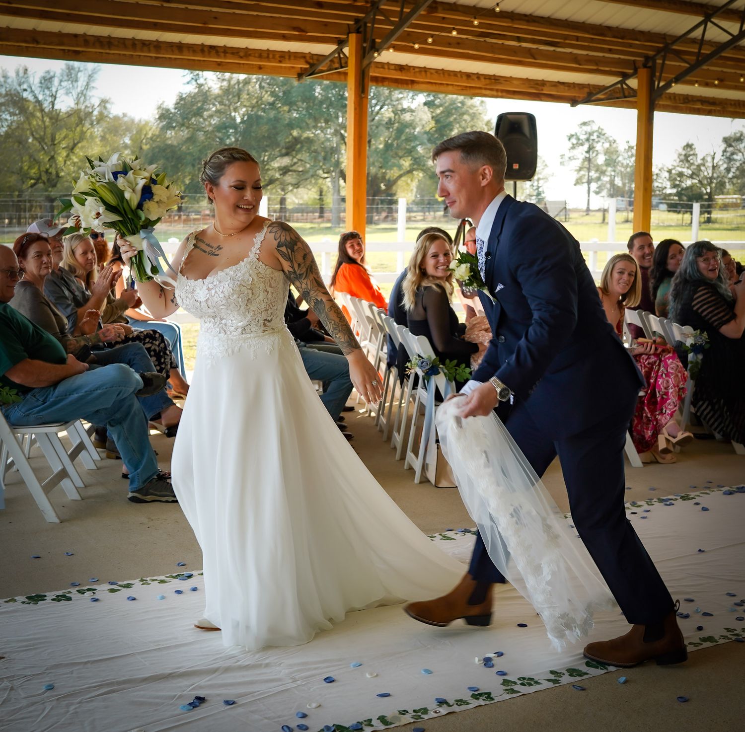 Smiling bride and groom celebrate walking back down aisle after outdoor wedding ceremony under wooden pavilion.