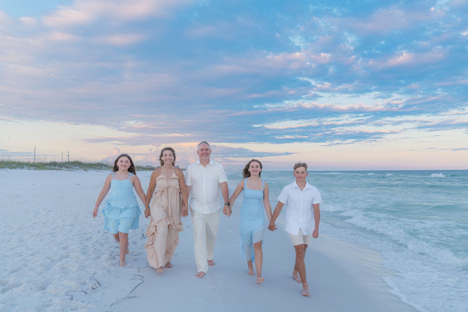 Family of five walking hand-in-hand on a white sand beach at sunset wearing coordinated pastel outfits.