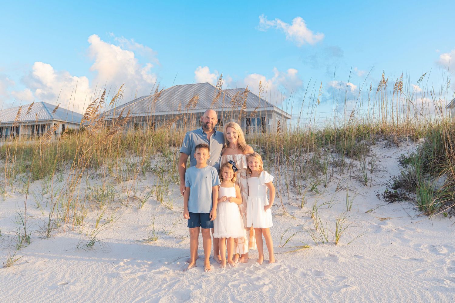 Family of five poses on white sand dunes with sea oats and beach house in background at sunset.