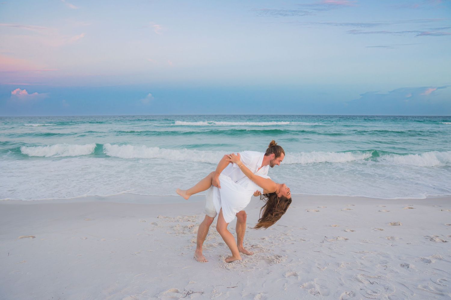 Couple dancing romantically on a white sand beach at sunset, waves and pastel sky in the background.