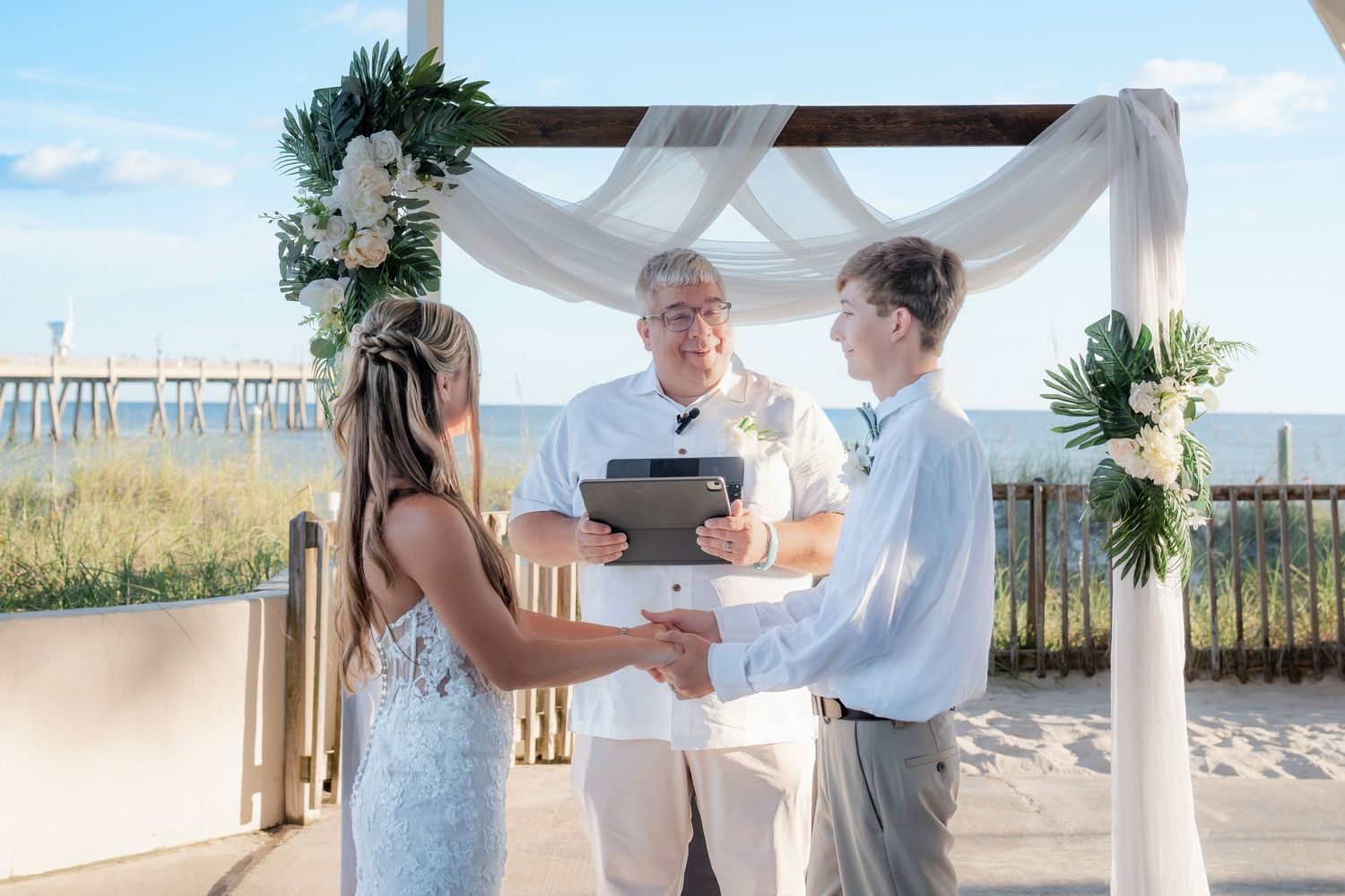 Beach wedding ceremony with couple exchanging vows under floral arch with ocean pier backdrop.