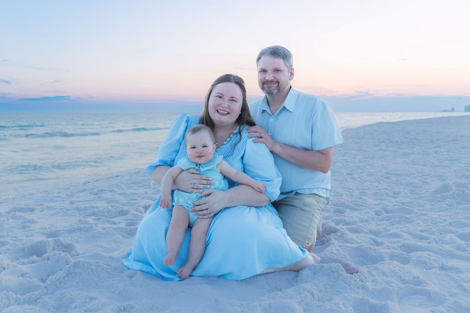 Family of three posing on a sandy beach at sunset, parents in light blue outfits holding a baby.