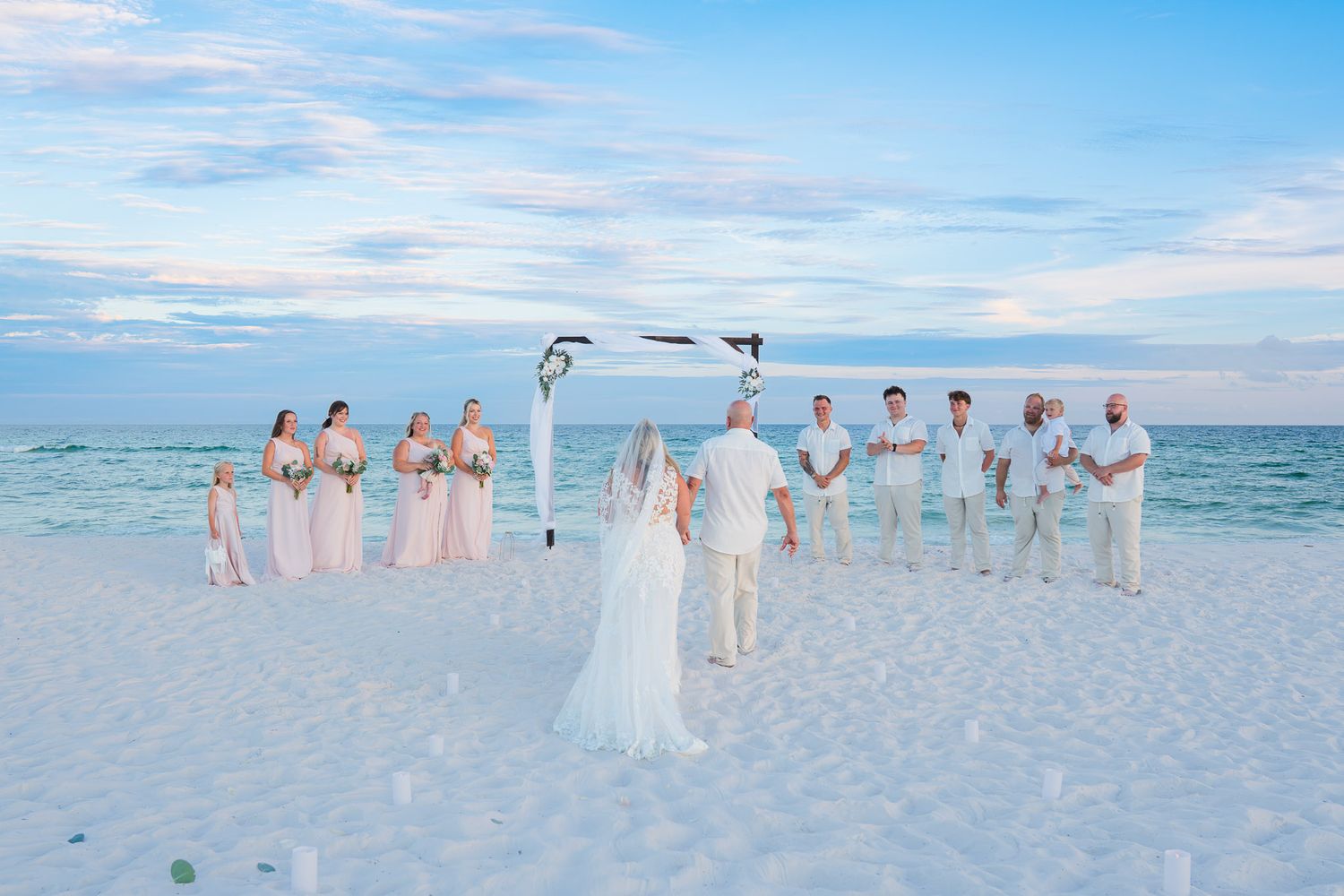 Beach wedding ceremony with bride, groom, wedding party in blush pink and tan attire, standing on white sand with ocean backdrop.