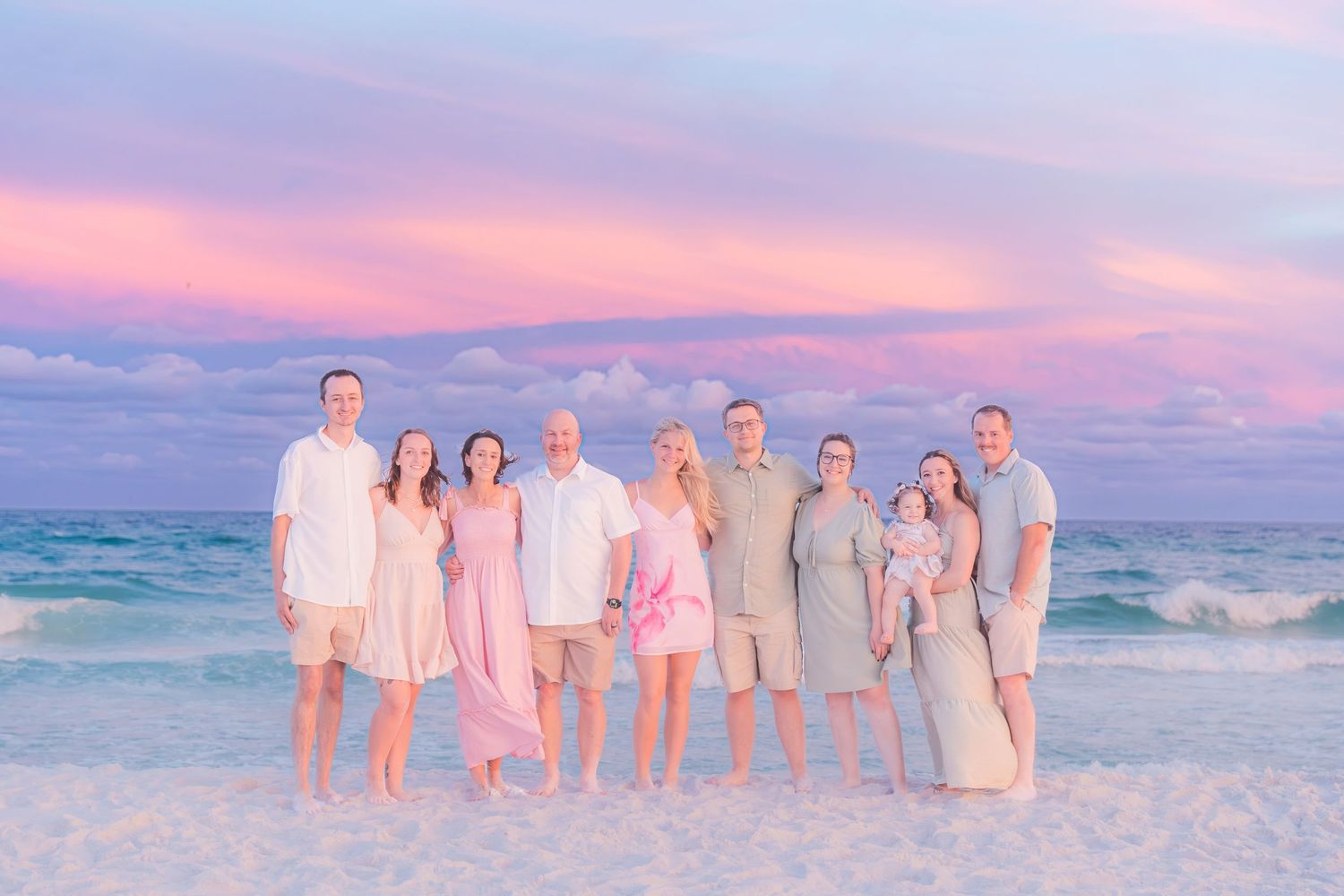 Extended family of nine stands together on a beach shoreline beneath a breathtaking pink sunset sky.