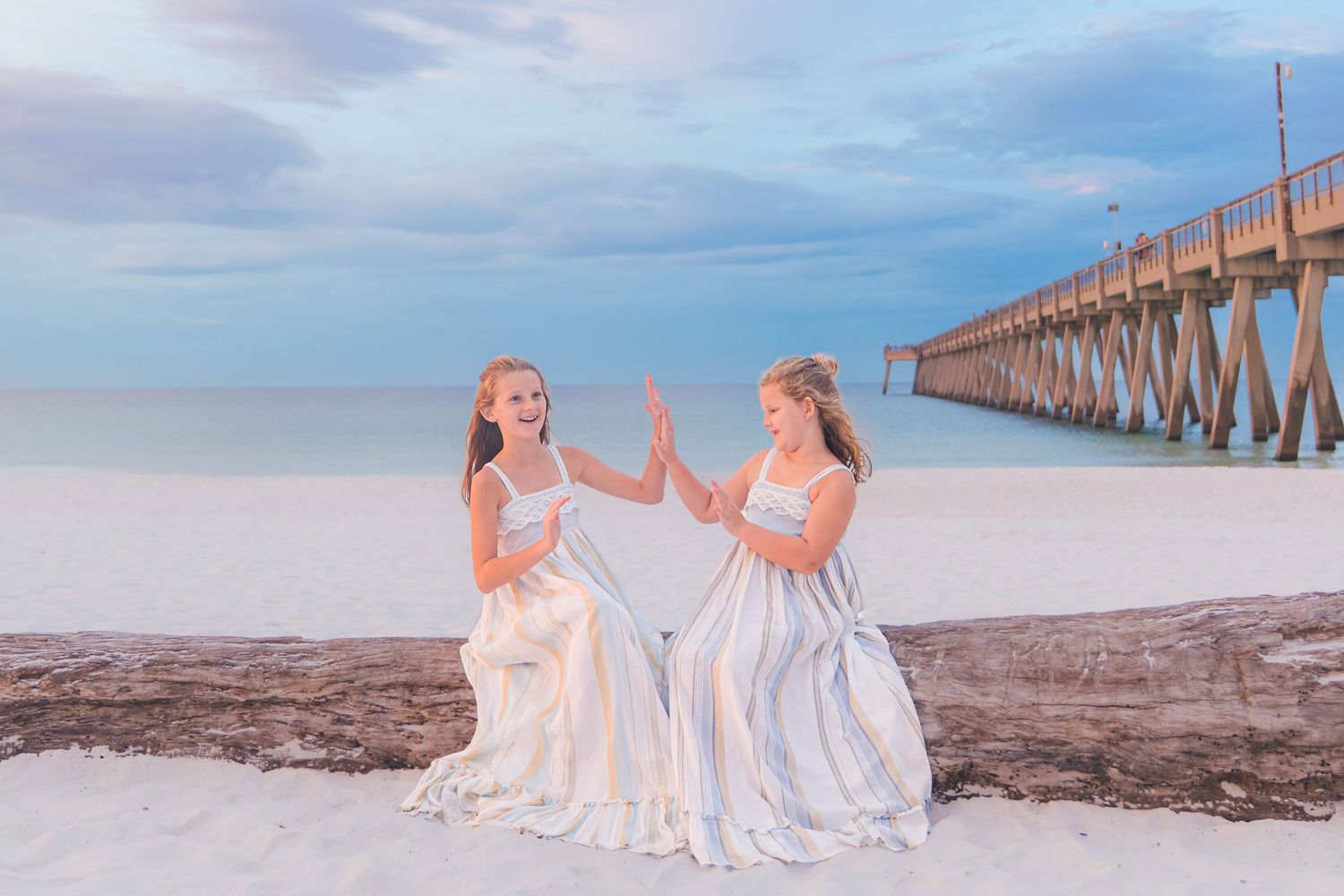 Two girls in white dresses high-fiving on a sandy beach near a pier at sunset.