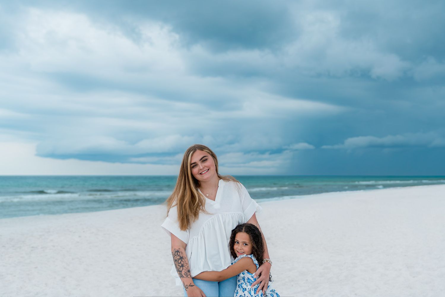 Woman and young girl smiling together on a white sand beach under dramatic cloudy skies.