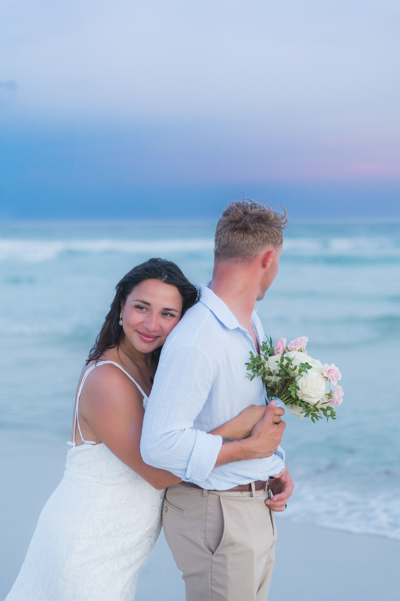 Bride hugging groom from behind on a beach at sunset, holding a bouquet of pink and white roses.