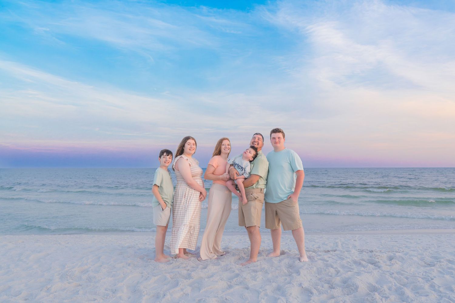 Family of six poses on a white sand beach at sunset with pastel sky backdrop.