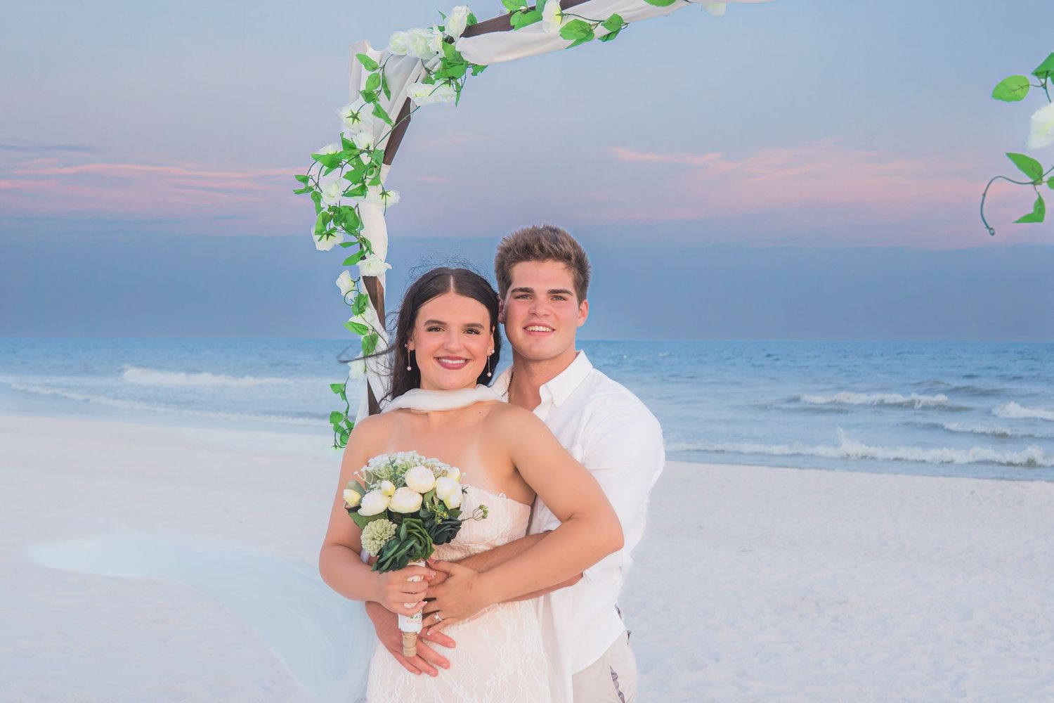 Couple in wedding attire posing on beach under floral arch with ocean sunset backdrop.