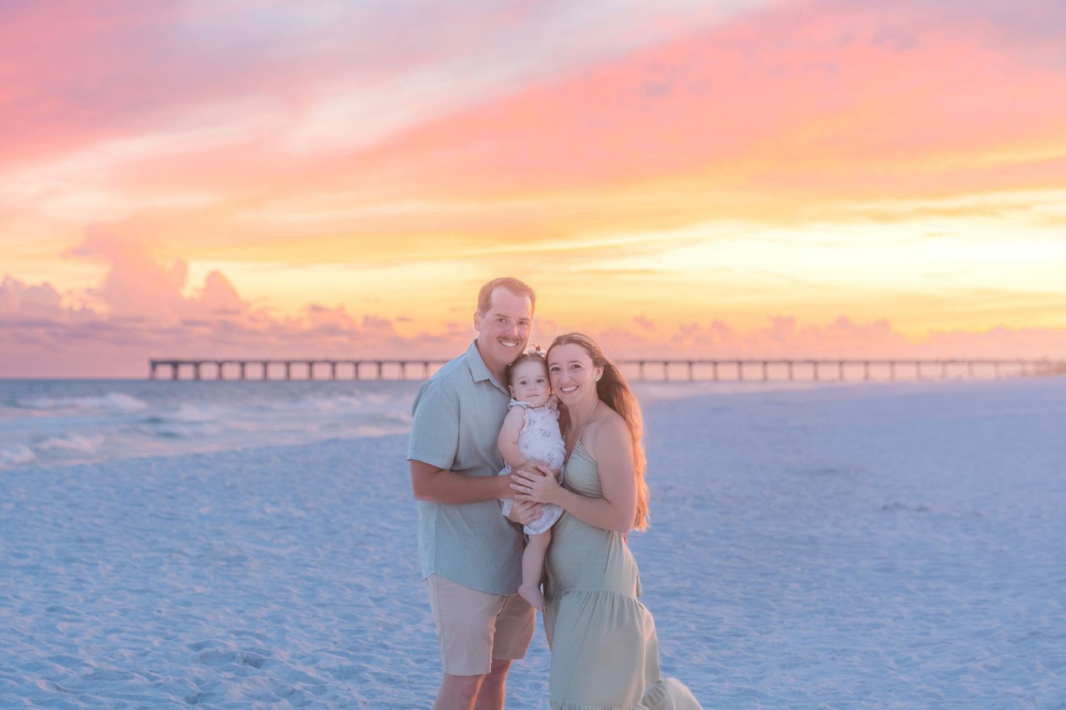 Family of three poses on a beach at stunning pink and orange sunset with a pier in the background.