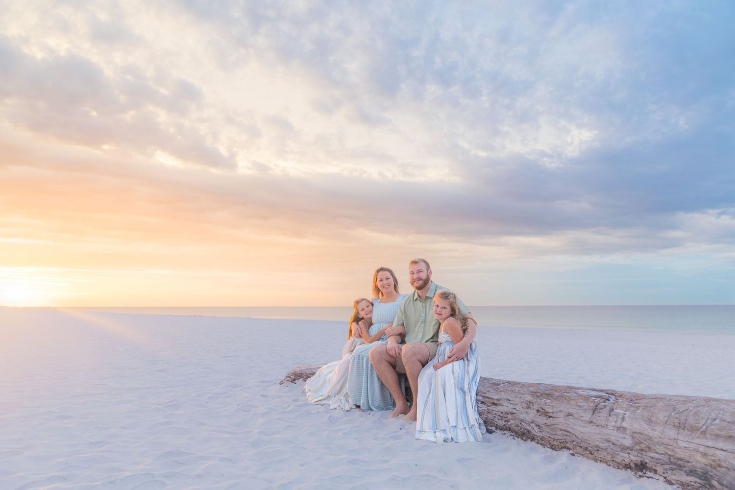 Family of four sitting on driftwood at a pastel sunset beach, white sand, soft sky colors.