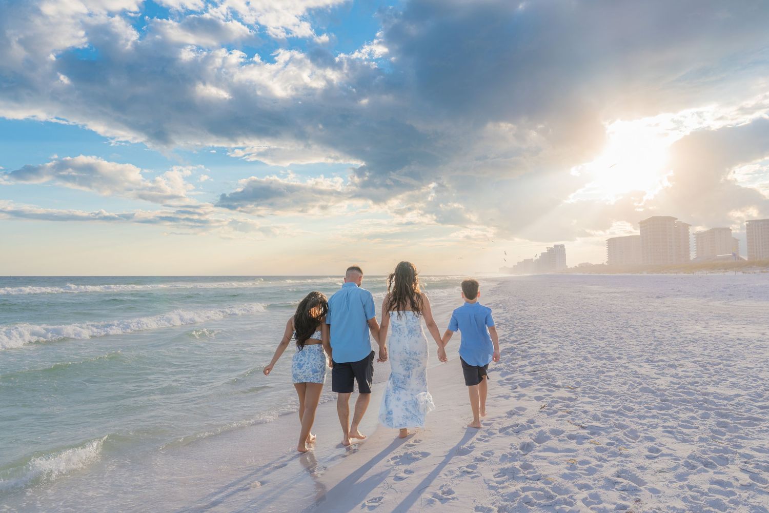 Family of four walking hand-in-hand on a sunny white sand beach with ocean waves and city skyline at sunset.