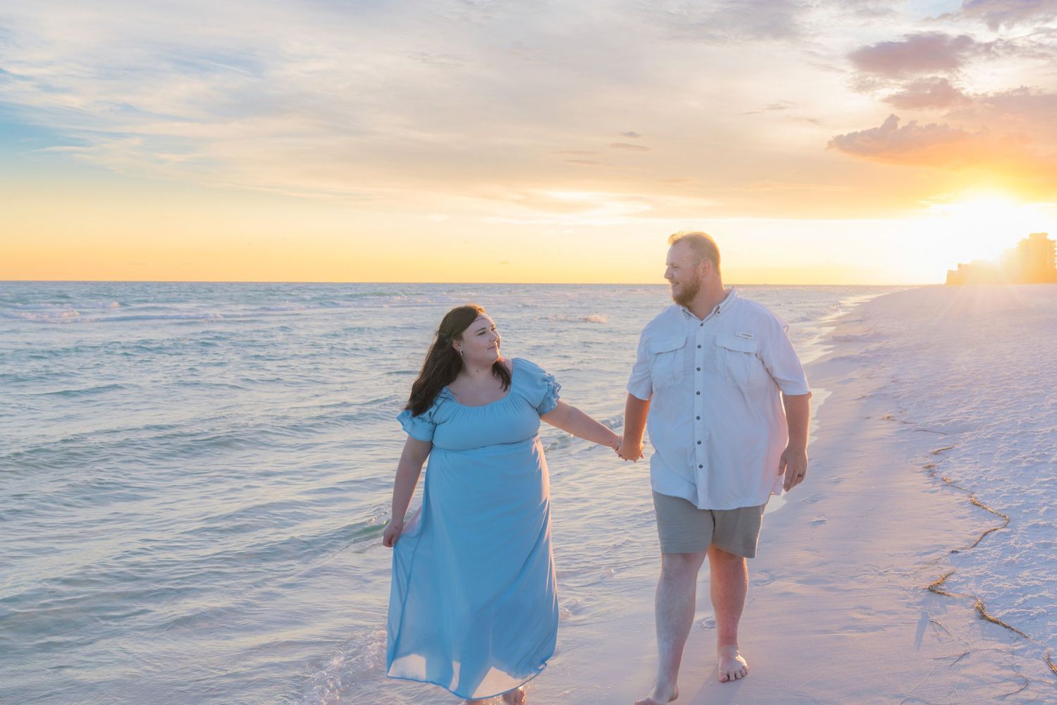 Couple holding hands walking on white sand beach at sunset, woman in blue dress, man in white shirt.