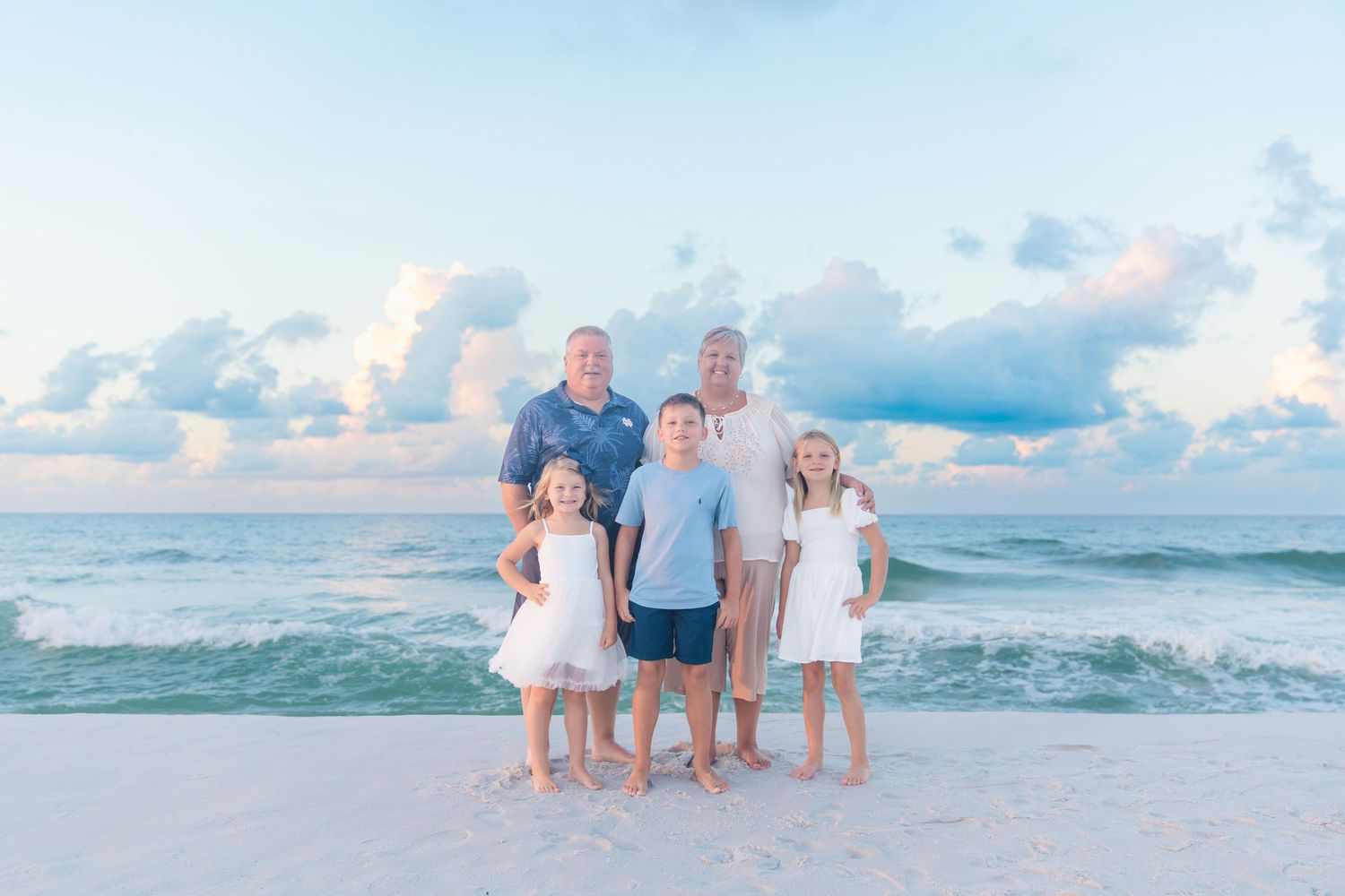 Family of five posing on a white sand beach at sunset with calm ocean waves and pastel sky behind them.