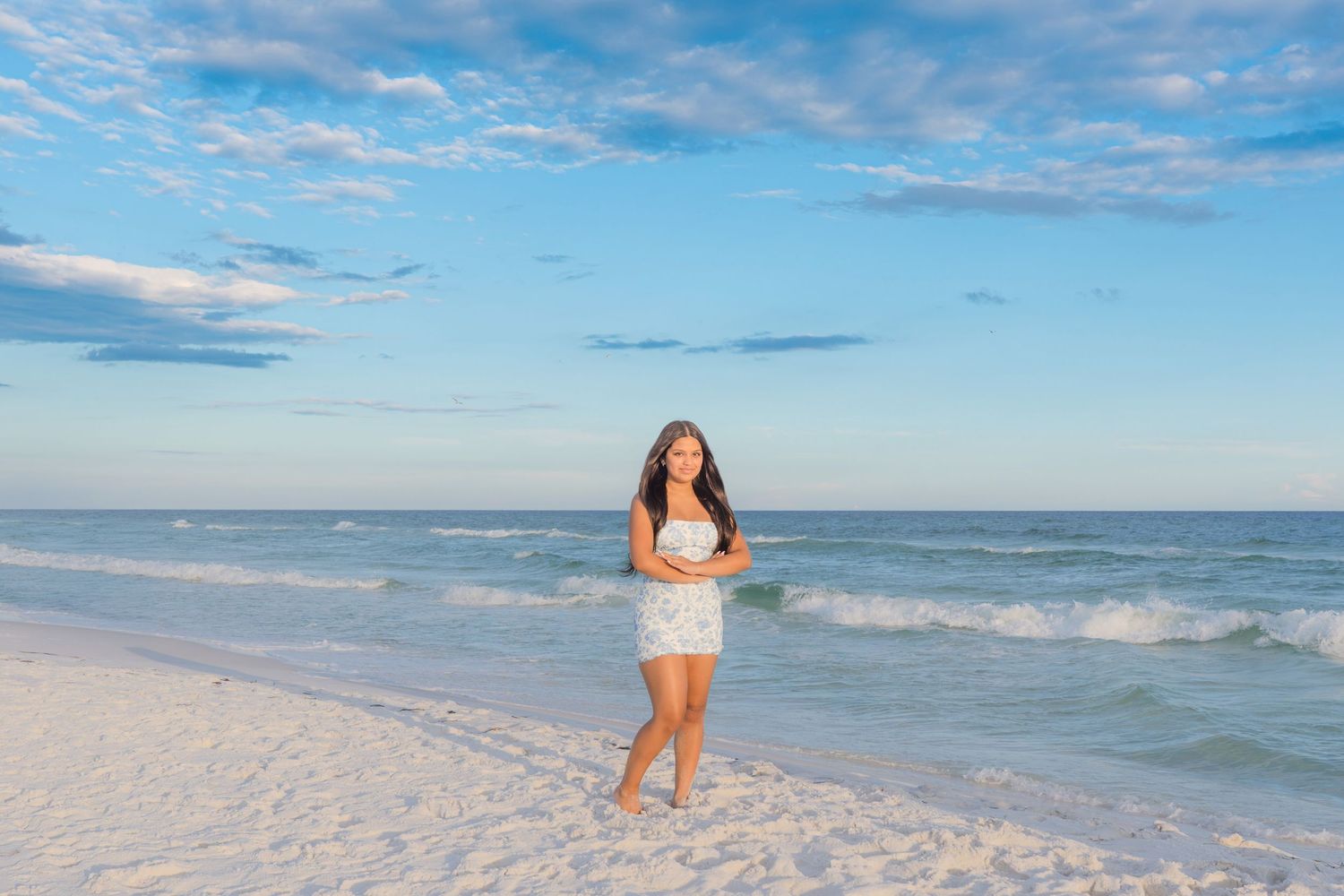 A young woman in a white dress walks on a pristine white sand beach with turquoise waves and a blue sky.