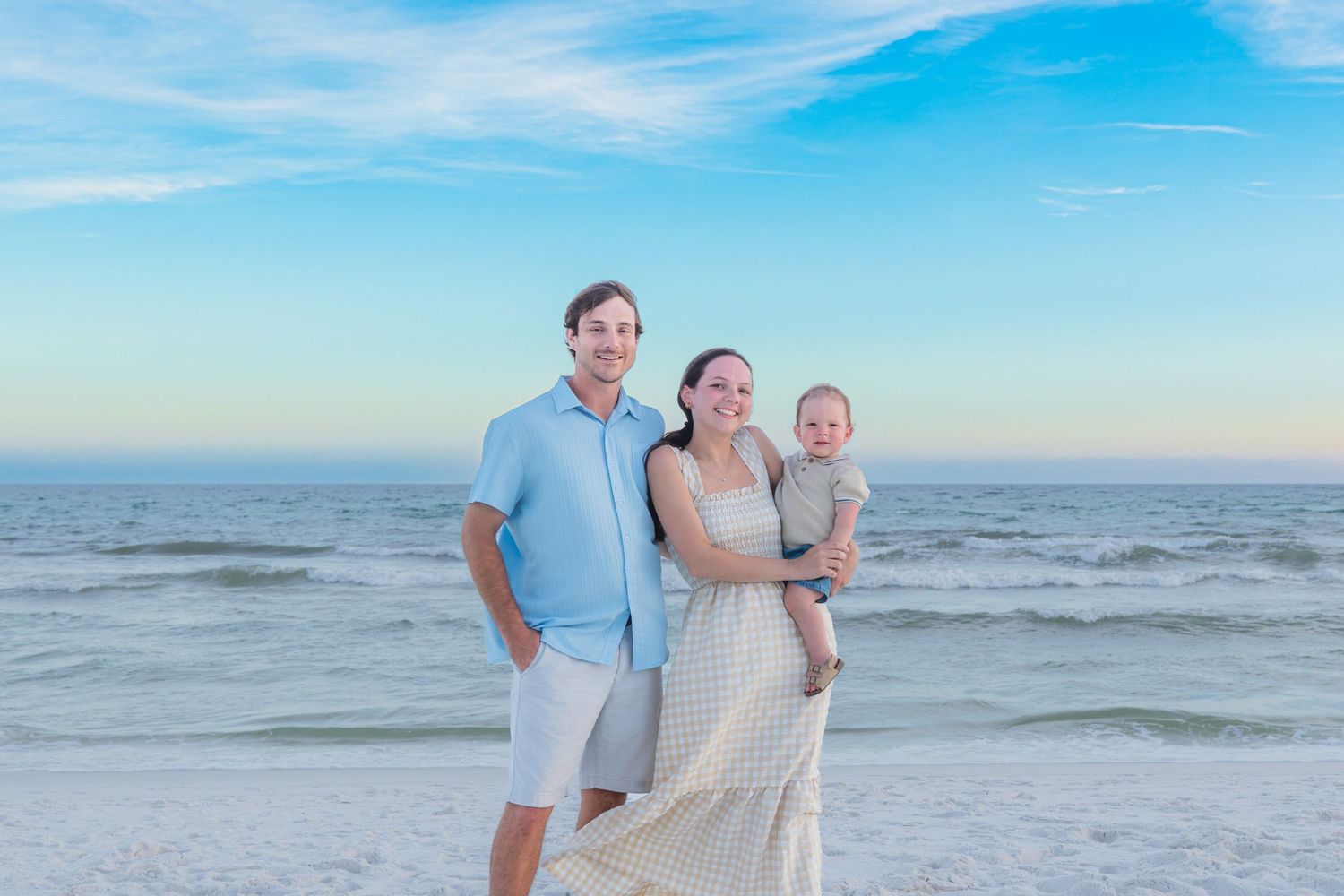 Young family of three posing on a beach at sunset with ocean behind them.